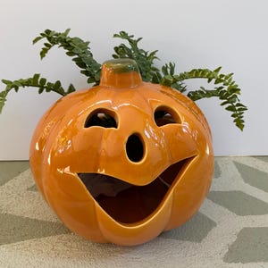 May include: A glossy orange ceramic pumpkin with a smiling face, perfect for Halloween decor. The pumpkin has cut-out eyes and a mouth, with green foliage sprouting from the top. A festive and decorative item.