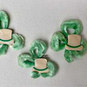 May include: Three green felt shamrock-shaped hair clips with a white felt top hat design. Each hat has a green felt band.
