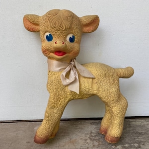 May include: Vintage yellow lamb toy with blue eyes, a red mouth, and a cream-colored ribbon tied around its neck. The lamb has textured fur and is approximately 10 inches tall.