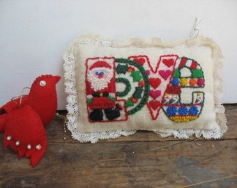 May include: A red felt bird ornament with pearl accents and a rectangular pillow ornament. The pillow features the word "LOVE" embroidered with Santa Claus, hearts, and colorful patterns. The pillow has a lace trim.