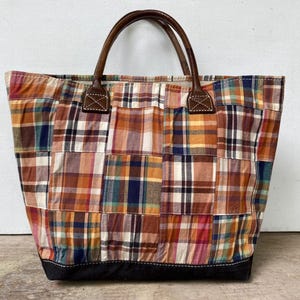 May include: A patchwork tote bag with a brown leather handle and base. The bag features a colorful plaid pattern in shades of orange, brown, blue, and white. The handles are attached with brown leather patches.
