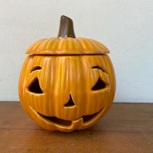 May include: A ceramic jack-o'-lantern container with a removable lid, featuring a glossy orange finish and a dark brown stem. The pumpkin has carved triangular eyes, a small triangular nose, and a wide, smiling mouth with a single tooth.