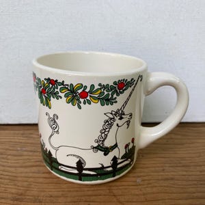 May include: A cream-colored ceramic mug featuring a black line drawing of a unicorn resting behind a green fence. The design includes a decorative garland of green leaves and red berries along the top rim, with the unicorn wearing a green collar.
