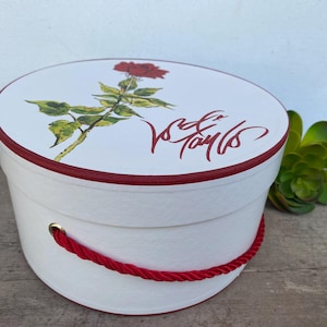 May include: A white round hat box with a red rose illustration on the lid. The box has a red trim and a red rope handle. The lid also features red script text.
