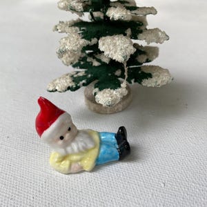 May include: A miniature Christmas scene featuring a small, ceramic gnome wearing a red hat, yellow shirt, and blue pants. A small, snow-covered Christmas tree is in the background. The gnome is lying down.