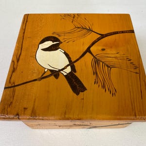 May include: A square wooden box with a bird and branch design. The bird is black and white, perched on a brown branch with brown leaves. The box is made of light brown wood.