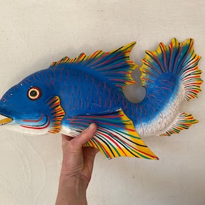 May include: A hand-painted, wooden fish sculpture. The fish is primarily blue with yellow, red, and green accents on the fins and tail. The fish has a detailed design and is held by a person's hand.