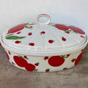 May include: White ceramic casserole dish with a lid, adorned with a vibrant red tomato pattern. The dish has a handle on the lid and two side handles, perfect for serving. The design features large and small tomatoes.