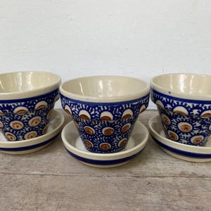 May include: Three ceramic flower pots with matching saucers. The pots are cream-colored inside and feature a blue and white floral pattern on the outside. The saucers are white with a blue stripe.