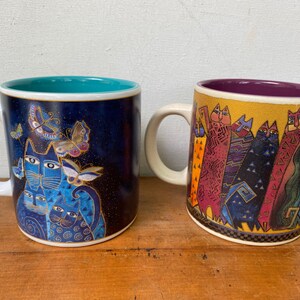 May include: Two ceramic mugs with white handles. The left mug features a dark blue background with blue cats and butterflies. The right mug has a yellow background with a row of colorful, patterned cats and a purple interior lining.