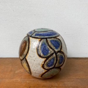 May include: A spherical ceramic object with a textured off-white base featuring a pattern of dark-outlined shapes in shades of blue, sage green, and brown. The surface has a glossy finish with visible glaze variations and dark, thick lines.