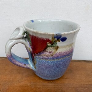 May include: A handmade ceramic mug with a unique handle design. The mug features a color palette of red, blue, purple, and white. The mug is sitting on a wooden surface.