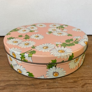 May include: A round, pink metal storage tin featuring a repeating pattern of white daisies with yellow centers and green leaves. The lid and the sides of the container are decorated with the same floral design, creating a cohesive look.