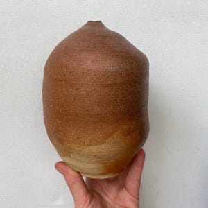 May include: A hand-thrown ceramic vase with a rounded, bulbous body and a narrow opening at the top. The surface features a textured, earthy terracotta brown finish that transitions into a lighter, sandy beige tone near the base.