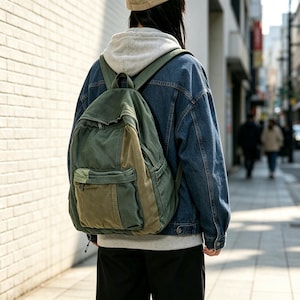 Könnte beinhalten: Ein zweifarbiger Rucksack aus olivgrünem und waldgrünem Canvas mit einem großen Hauptfach mit Reißverschluss und einer kleineren Vordertasche. Der Rucksack verfügt über verstellbare Schultergurte und wird über einem grauen Kapuzenpullover getragen.