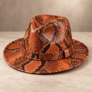 Pode incluir: Um chapéu fedora com um padrão de pele de cobra laranja e preto proeminente na copa e na aba larga. O chapéu inclui uma faixa de pele de cobra a condizer que envolve a base da copa, criando uma aparência coesa e texturizada.