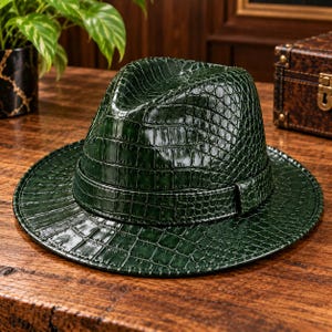 May include: A dark green fedora hat featuring a glossy, embossed crocodile skin texture throughout the crown and brim. The hat includes a matching green band wrapped around the base of the crown, showcasing a consistent scale pattern.