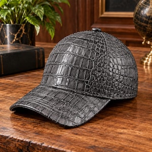 May include: A baseball cap featuring a dark grey, textured finish that mimics the appearance of crocodile skin. The cap has a structured crown and a curved brim, with visible stitching along the panels and edges of the material throughout.