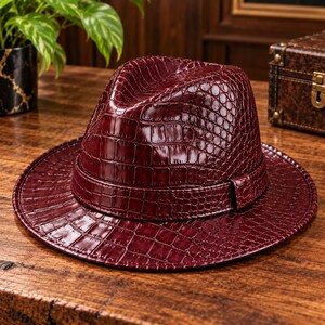 Peut inclure: Un chapeau fedora bordeaux profond doté d'une texture brillante en relief façon peau de crocodile sur toute la calotte et le large bord. Le chapeau comprend une bande ton sur ton assortie autour de la base, affichant un motif reptilien.