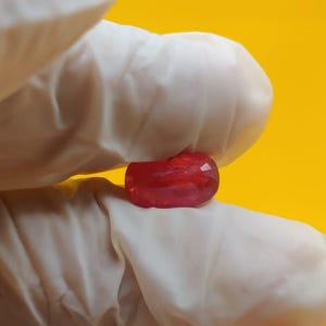 以下が含まれることがあります： ファセットカットが施された、小さく楕円形の半透明の赤い宝石。深いクリムゾンカラーの石が、白いラテックス手袋をはめた2本の指の間に挟まれており、背景は無地の明るい黄色です。