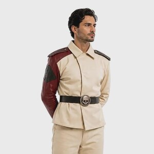 Op de afbeelding: Een beige jas met een puntige kraag, donkerrode mouwen en schouderstukken, gecombineerd met een bijpassende beige broek. Een zwarte riem met een opvallende zilverkleurige schedelgesp wordt in de taille gedragen. Beige veterschoenen maken de outfit af.