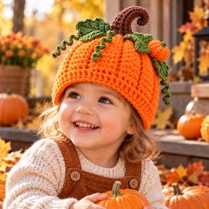 May include: A bright orange crochet pumpkin hat featuring a dark brown curly stem, green crocheted leaves, and a small orange pumpkin accent on the side. The hat is textured with a ribbed stitch pattern and is worn by a smiling child in a sweater.