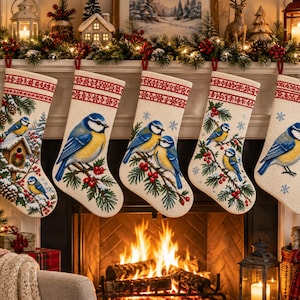 May include: Five cream-colored Christmas stockings hanging in a row, each featuring a red patterned cuff and embroidered designs of blue and yellow birds perched on pine branches with red berries and snow-dusted evergreen foliage.