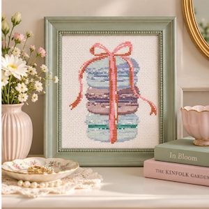 May include: A framed cross-stitch embroidery featuring a stack of three pastel-colored macarons tied with a pink ribbon. The artwork is displayed in a sage green frame with a beaded inner edge, showcasing intricate needlework on white fabric.