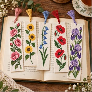 Könnte beinhalten: Fünf rechteckige Lesezeichen aus Stoff mit Kreuzstich-Blumenmustern aus rosa Rosen, gelben Sonnenblumen, blauen Glockenblumen, roten Mohnblumen und violetten Schwertlilien, jeweils mit einer farblich passenden Quaste versehen.