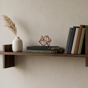 Può includere: Una mensola da parete in legno marrone scuro con due supporti verticali. Sulla mensola si trovano un vaso in ceramica bianca contenente un pennacchio di erba della pampa essiccata, una scultura geometrica in filo di rame e cinque libri colorati.