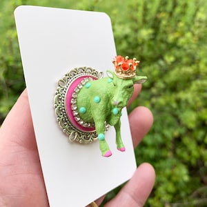 May include: A brooch featuring a lime green cow figure with turquoise polka dots and pink hooves, wearing a small orange crown. The cow is mounted on a circular silver-toned frame with a hot pink centre and a border of clear rhinestones.