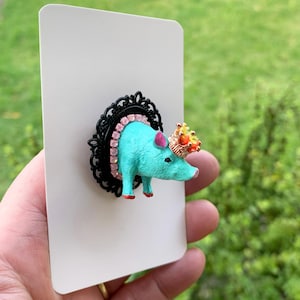 May include: A turquoise pig figurine brooch mounted on a black ornate oval frame. The pig features a gold-toned crown with orange and yellow accents, pink inner ears, and red hooves. A border of small pink rhinestones encircles the pig's neck.