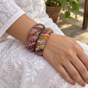 May include: Two thick bangle bracelets featuring a vibrant, multi-colored marbled pattern with wavy, repeating lines in shades of pink, yellow, green, orange, and blue. The bracelets have a smooth, rounded exterior surface and a polished finish.