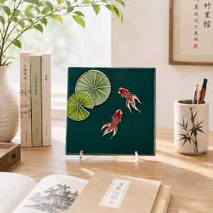 May include: A square, dark green ceramic tile featuring two raised, red and white goldfish figures and two light green, textured lily pads. The tile is displayed on a clear acrylic stand, showcasing a detailed relief design of aquatic life.