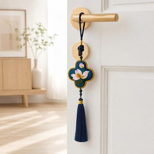 Puede incluir: Un adorno colgante decorativo con un colgante de tela azul verdoso de cuatro lóbulos, bordado con flores de loto rosas y blancas, hojas verdes y un borde dorado. Incluye un cordón azul oscuro, pequeñas cuentas verdes y una borla larga azul oscuro.
