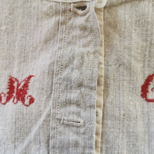 Może przedstawiać: Zbliżenie na lnianą koszulę z czerwonymi haftowanymi inicjałami "M" i "C" z przodu. Koszula zapinana jest na guziki i ma prosty design.