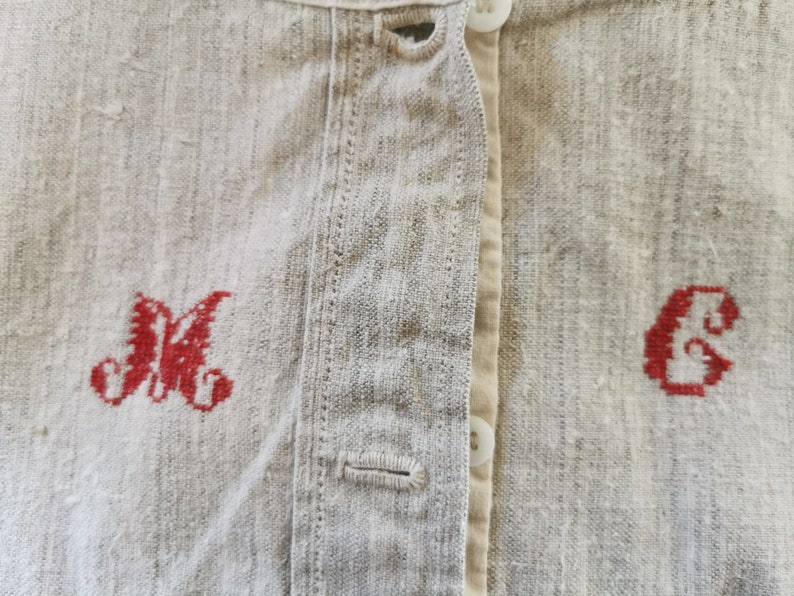Może przedstawiać: Zbliżenie na lnianą koszulę z czerwonymi haftowanymi inicjałami "M" i "C" z przodu. Koszula zapinana jest na guziki i ma prosty design.