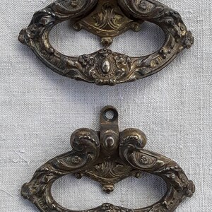 May include: Two antique bronze drawer pulls with ornate, scrollwork designs. Each pull features a curved handle with decorative embellishments and a central oval medallion. The pulls are mounted on a light-colored fabric background.