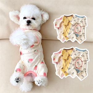 May include: A white dog wearing a cream-colored pet jumpsuit with a pink strawberry print and pink trim. Beside the dog are two additional sets of pet jumpsuits in three colorways: pink with strawberries, yellow with brown accents, and cream with blue trim and smiley faces.