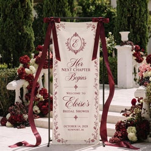 Op de afbeelding: Een verticaal crèmekleurig stoffen spandoek aan een zwarte metalen standaard, voorzien van donkerrode bloemenranden en de tekst Her NEXT CHAPTER Begins WELCOME TO Eloise's BRIDAL SHOWER OCTOBER 14, 2028 NEWPORT, RI, afgewerkt met satijnen linten.