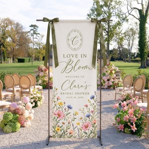May include: A vertical cream-colored fabric banner suspended from a dark metal stand with olive green ribbon bows. The banner features a floral border at the bottom and text reading: Love in Bloom, Welcome to Clara's Bridal Shower, June 18, 2028.
