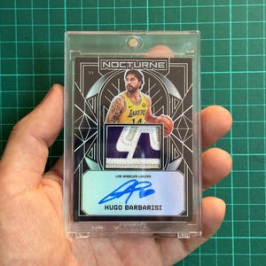 Könnte beinhalten: Eine Sport-Sammelkarte in einer durchsichtigen magnetischen Kunststoffhülle. Die Karte zeigt einen Basketballspieler im gelben Trikot der Los Angeles Lakers, einen Stoffaufnäher, den Text NOCTURNE, 1/1 und ein blaues Autogramm von Hugo Barbarisi.