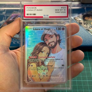 Peut inclure: Une carte à collectionner Pokemon personnalisée intitulée Leana et Hugo, notée GEM MT 10, scellée dans un boîtier en plastique transparent. La carte présente une illustration style anime d'un couple, du texte français et un fini holographique.