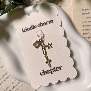 May include: A gold-toned metal charm featuring a small book, a star, and a sword attached to a paperclip-style link. The charm is mounted on a scalloped-edge white card printed with the text kindle charm and chapter nineteen in black font.