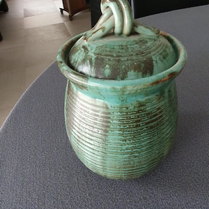 Op de afbeelding: Een keramieken pot met deksel voorzien van een geknoopte handgreep. De pot heeft een ronde vorm met horizontale ribbeltexturen en een glazuur in tinten turquoise, petrol en donkerbruin. Het deksel heeft hetzelfde kleurenpalet en afwerking.