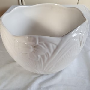Op de afbeelding: Een glanzende witte keramische kom met een geschulpte rand en een reliëfpatroon van bloemmotieven en bladeren die rondom de buitenkant zijn aangebracht. De kom heeft een brede, ronde vorm met een gladde, reflecterende afwerking.