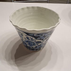 Op de afbeelding: Een keramische plantenpot met een geschulpte rand en een witte binnenkant. De buitenkant is versierd met een blauw-wit bloemmotief, inclusief gedetailleerde bloesems en bladeren in een herhalend patroon rondom de taps toelopende vorm.