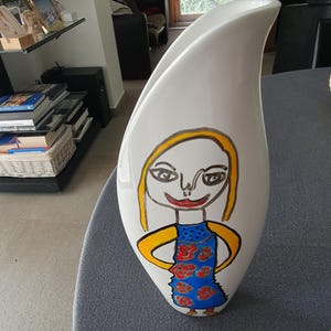 Könnte beinhalten: Eine hohe, weiße, geschwungene Keramikvase mit einer handgemalten Illustration einer Figur mit gelben Haaren, einem blauen Kleid mit roten Blumenmustern und schwarzen Umrissen. Die Vase hat eine glatte, glänzende Oberfläche und einen asymmetrischen Rand.