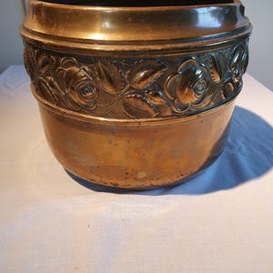 Könnte beinhalten: Ein runder, kupferfarbener Metallübertopf mit einem breiten, erhabenen Band am oberen Rand. Dieses Band ist mit einem durchgehenden Reliefmuster aus blühenden Rosen und detaillierten Blättern verziert. Die Oberfläche weist eine natürliche Patina auf.