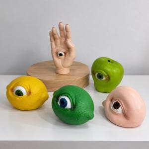 May include: A collection of five surreal sculptures featuring a pale hand with an eye in the palm, a bright yellow lemon with an eye, a vibrant green lime with an eye, a green apple with an eye, and a smooth pale pink rounded form with an eye.